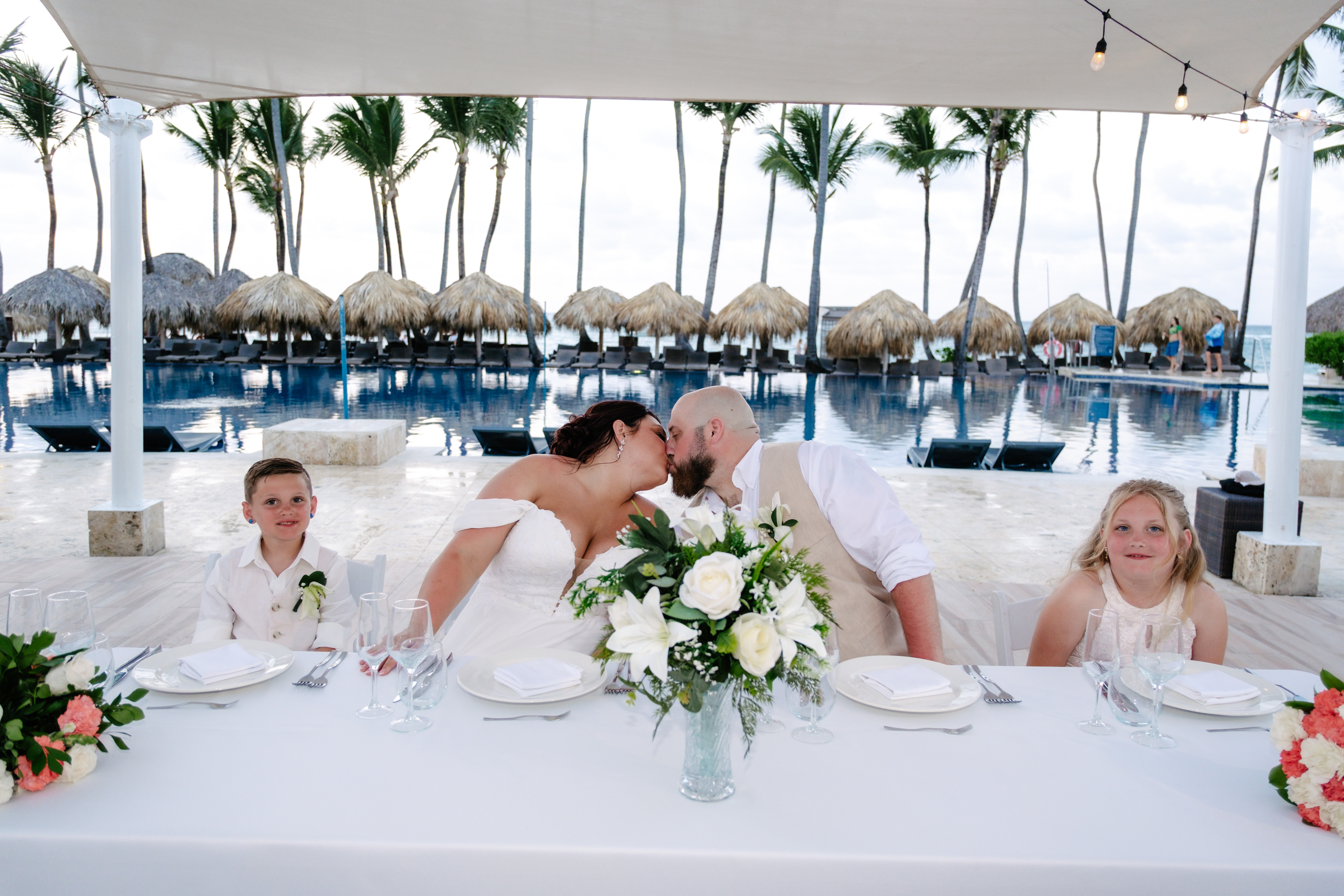Kelsie and Matthew share a kiss at their destination wedding, seated at a poolside table with their two children by their side.