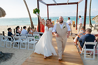 Kelsie and Matthew walk hand in hand down a wooden aisle on the sand, surrounded by guests seated on either side, as their beautiful destination wedding unfolds by the beach.