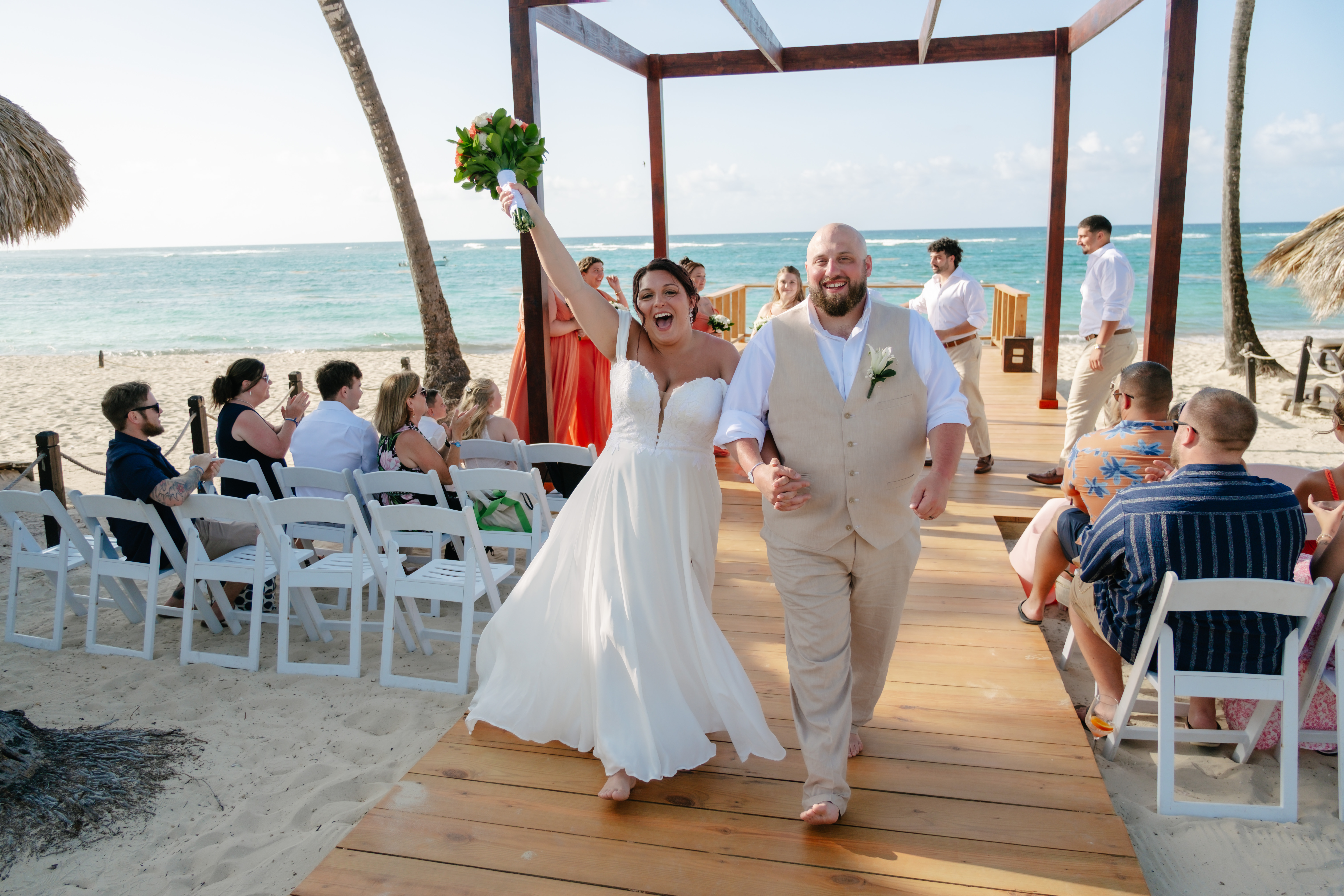 Kelsie and Matthew walk hand in hand down a wooden aisle on the sand, surrounded by guests seated on either side, as their beautiful destination wedding unfolds by the beach.
