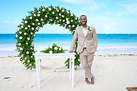 A man in a beige suit stands barefoot beneath a floral arch on the beach, ready for a beautiful destination wedding.