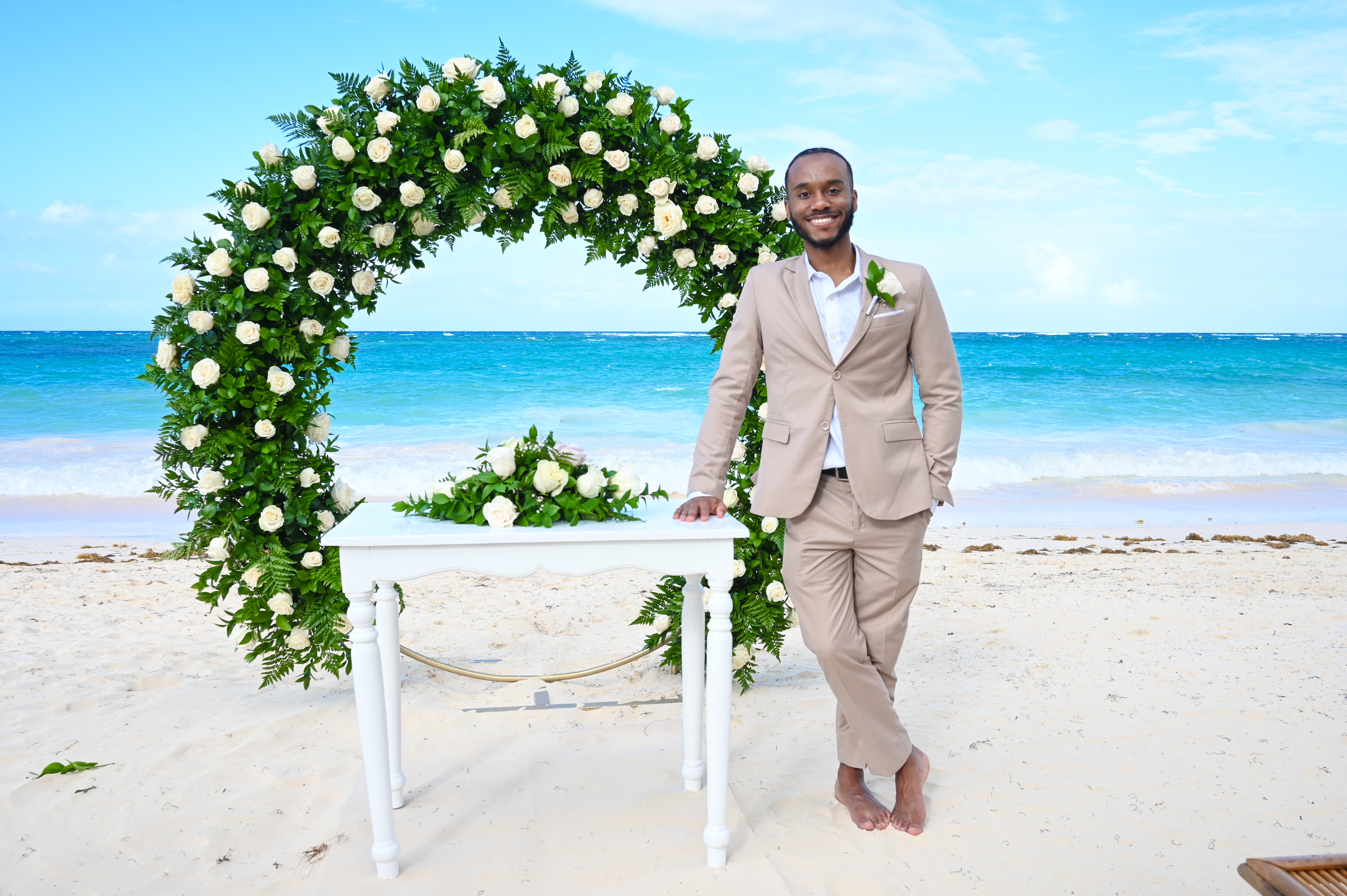 A man in a beige suit stands barefoot beneath a floral arch on the beach, ready for a beautiful destination wedding.