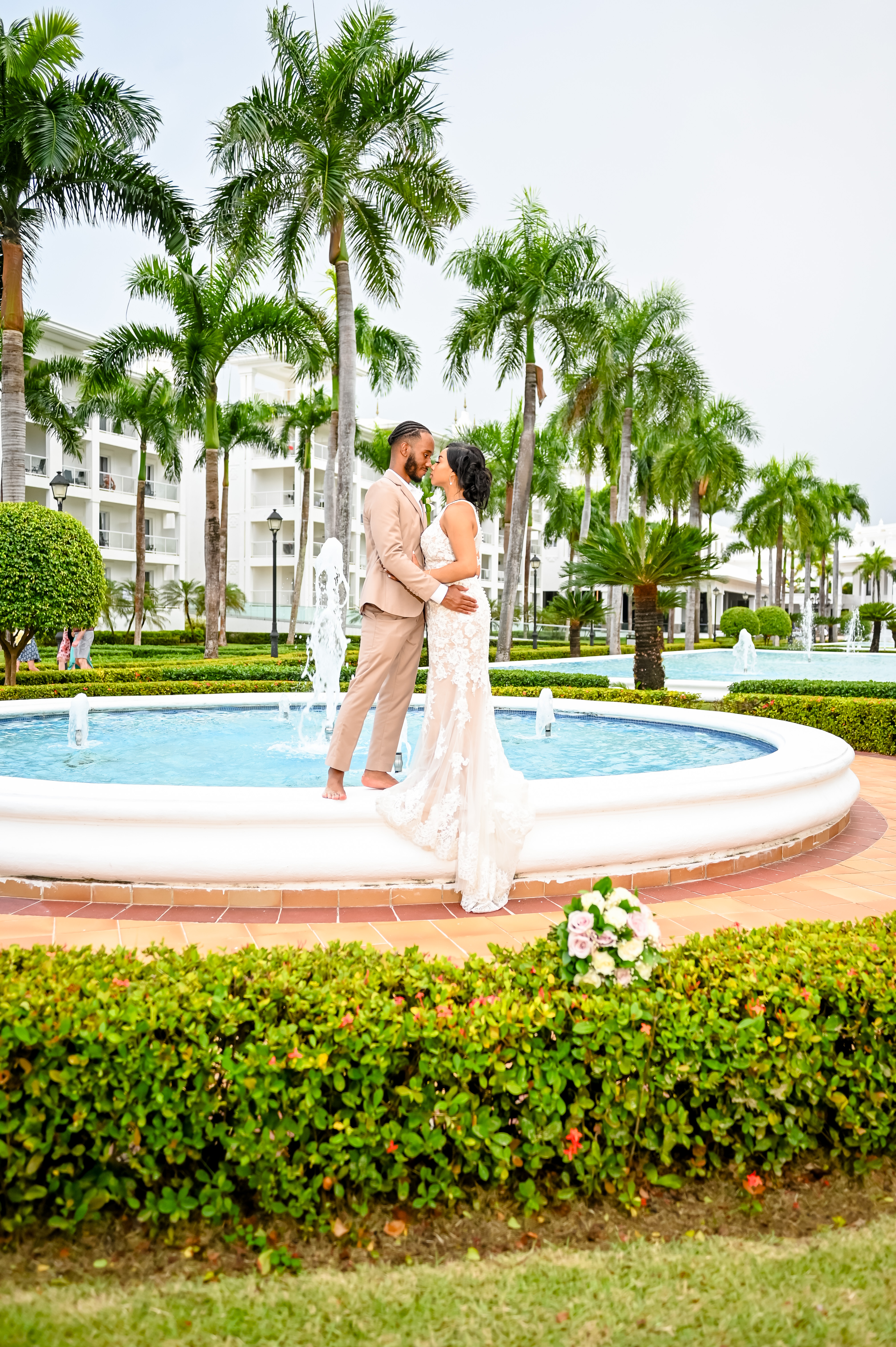 Trey and Kimberly pose in elegant wedding attire by a fountain surrounded by palm trees, capturing the essence of a destination wedding.