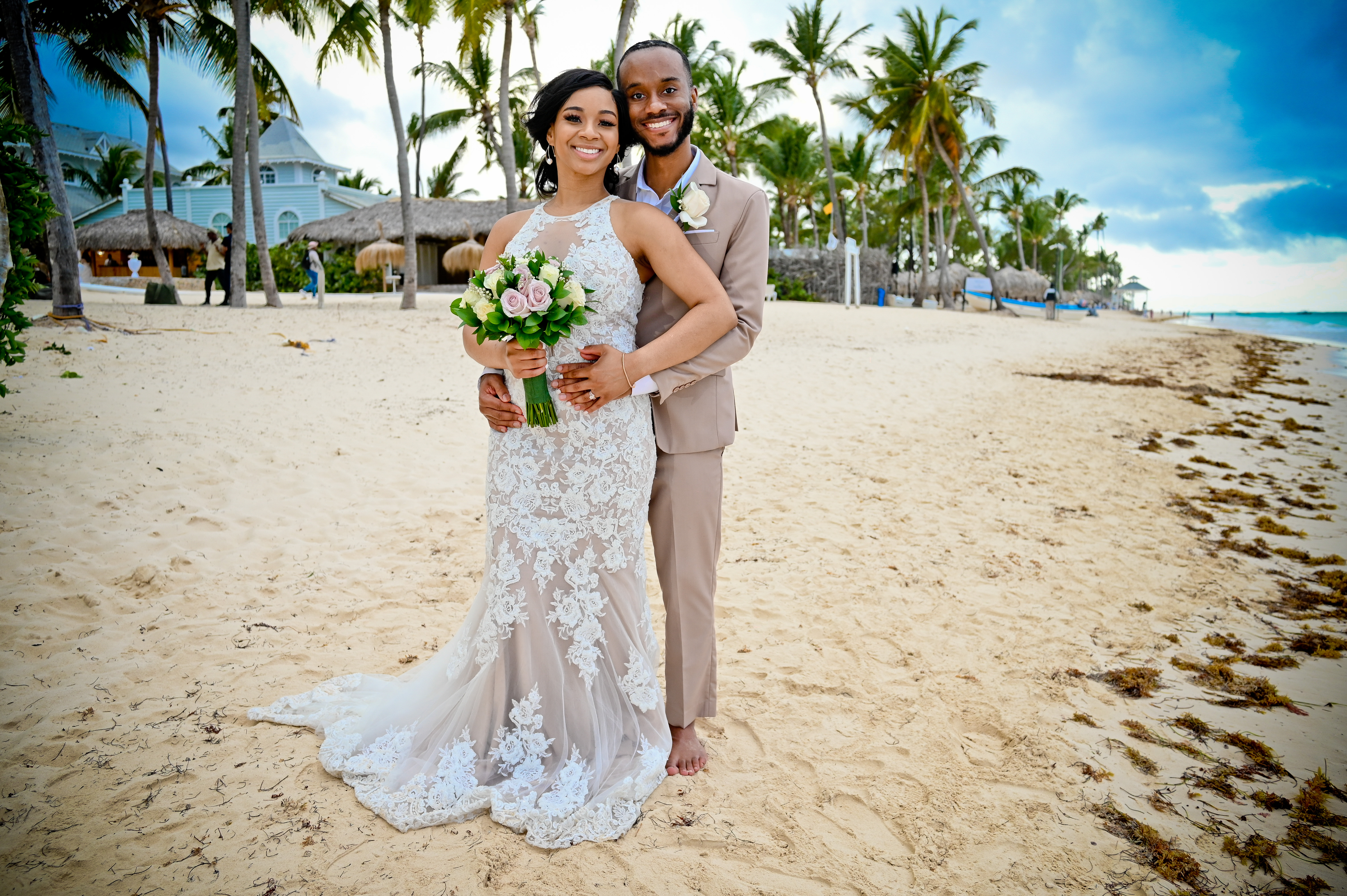 Trey and Kimberly pose together on a sandy beach lined with palm trees, capturing a beautiful moment from their destination wedding.