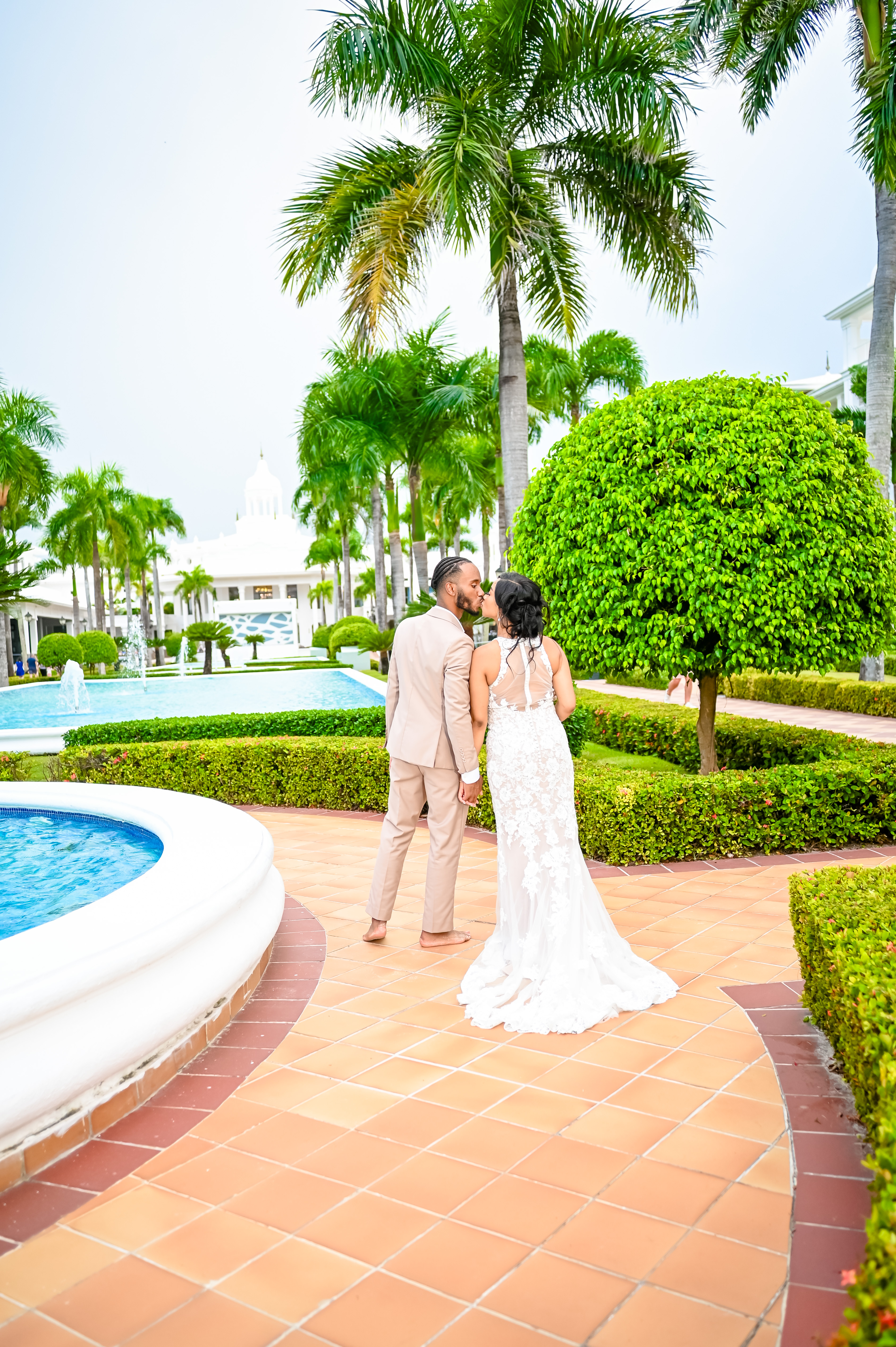 Trey and Kimberly stroll along a tiled pathway by the pool, surrounded by palm trees and lush gardens at their destination wedding.