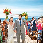 The bride and groom walk down the aisle on a beautiful beach as guests applaud and celebrate their destination wedding.