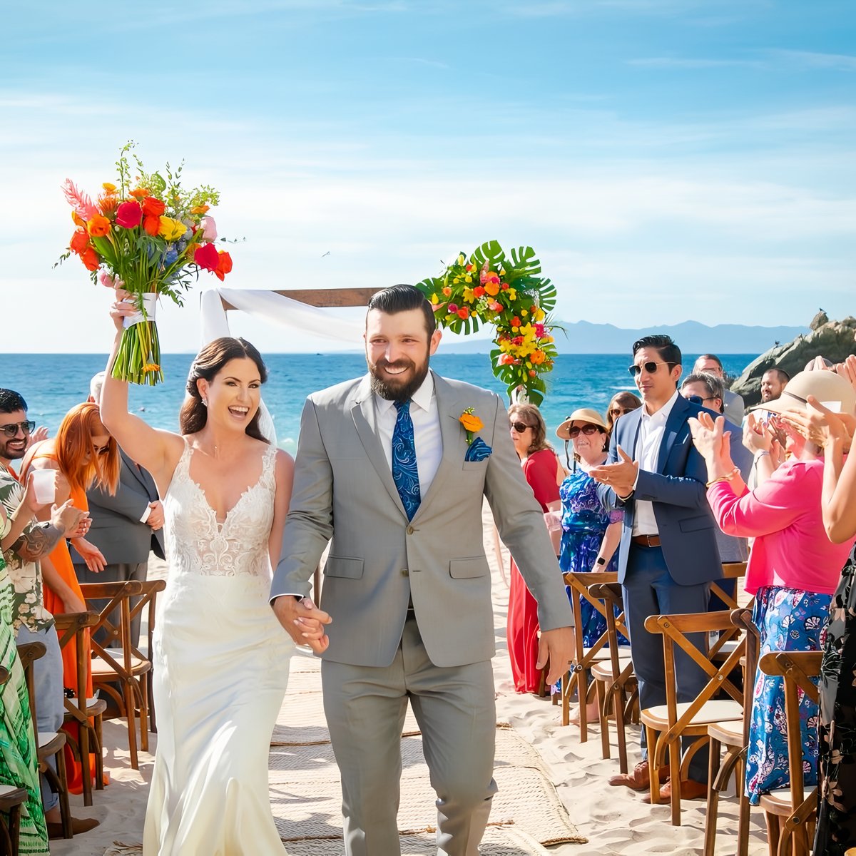 The bride and groom walk down the aisle on a beautiful beach as guests applaud and celebrate their destination wedding.