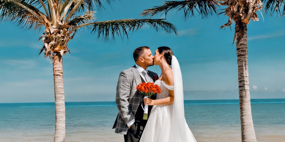At their destination wedding at Moon Palace Nizuc, the bride and groom share a kiss on the beach, with the bride holding a bouquet of red roses.