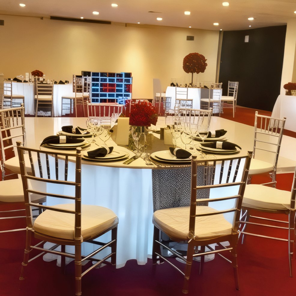 A round table elegantly set for a destination wedding, featuring plates, glasses, black napkins, and a striking red flower centerpiece in a beautifully decorated event hall.