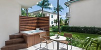 Ocean Maya Royale patio: An idyllic spot for a destination wedding, featuring a romantic hot tub, two inviting chairs, a table for intimate moments, lush greenery all around, and scenic nearby buildings to complete the perfect setting.