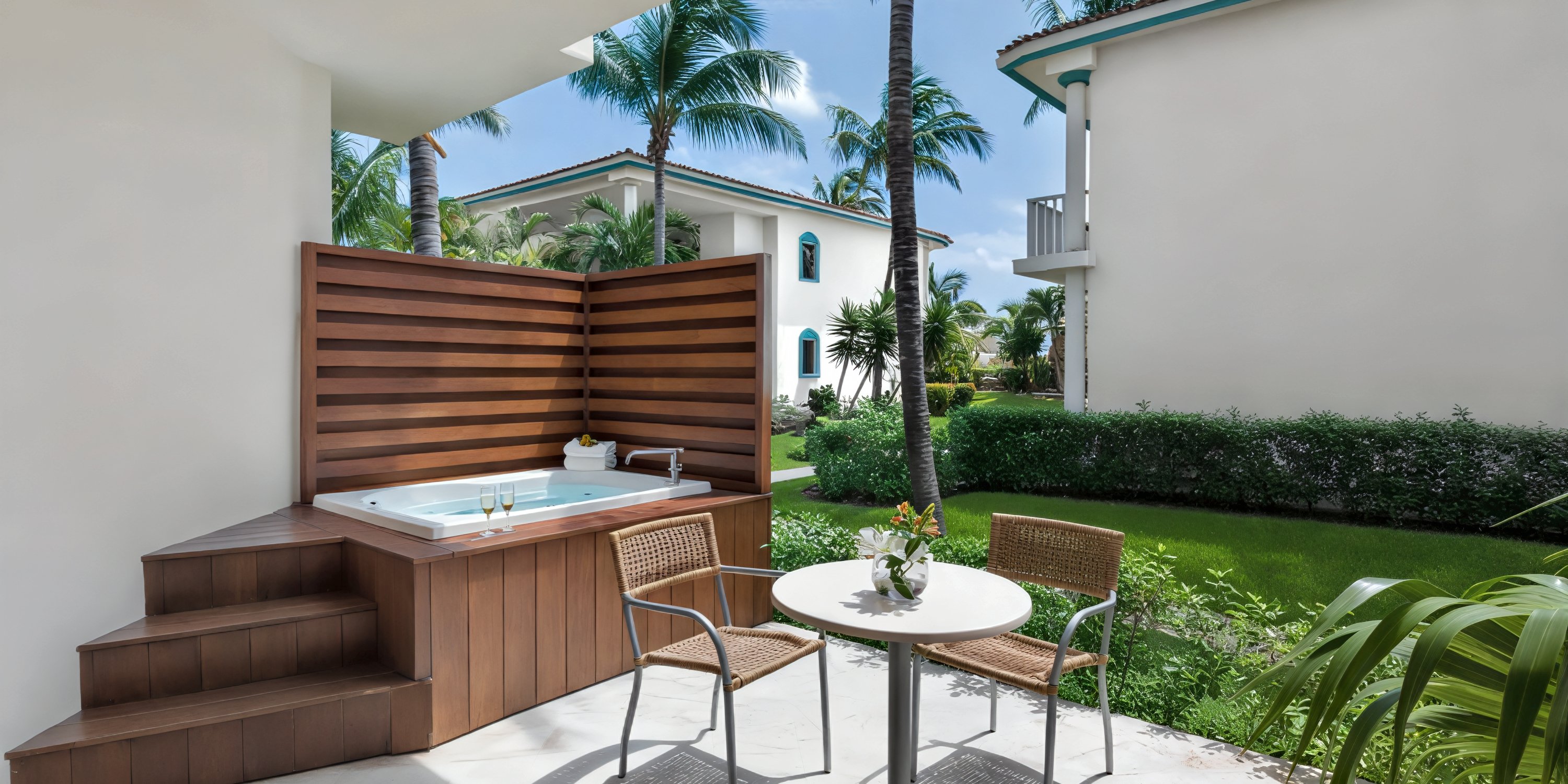 Ocean Maya Royale patio: An idyllic spot for a destination wedding, featuring a romantic hot tub, two inviting chairs, a table for intimate moments, lush greenery all around, and scenic nearby buildings to complete the perfect setting.