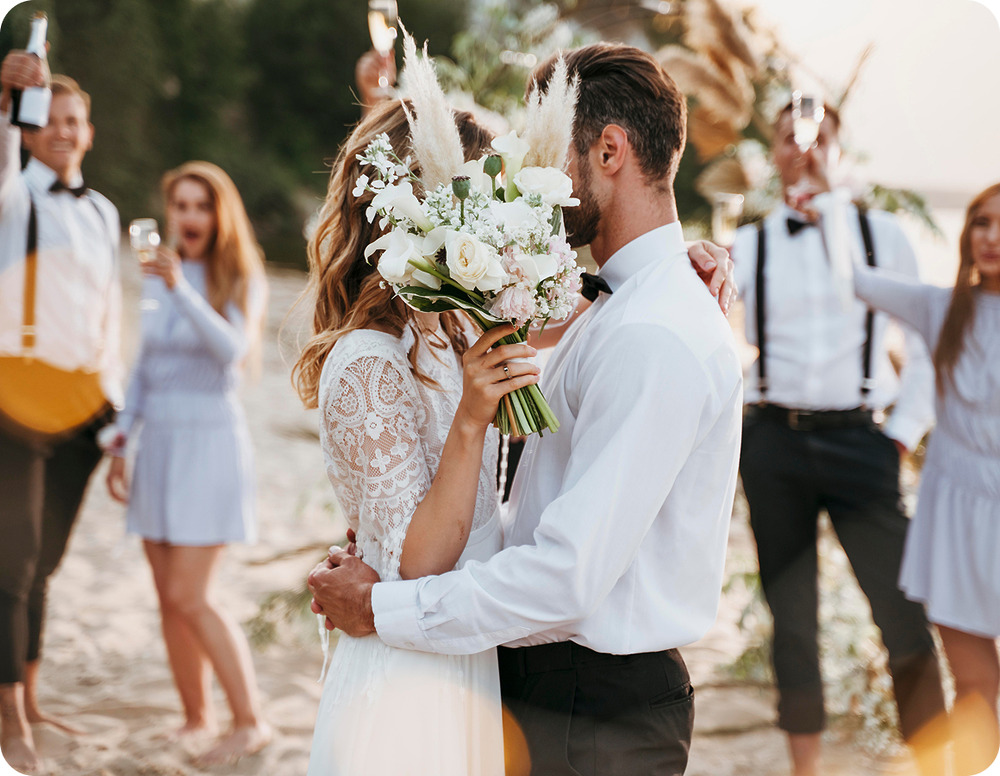 The bride and groom embrace on the beach during their destination wedding, holding flowers while friends celebrate joyfully in the background.