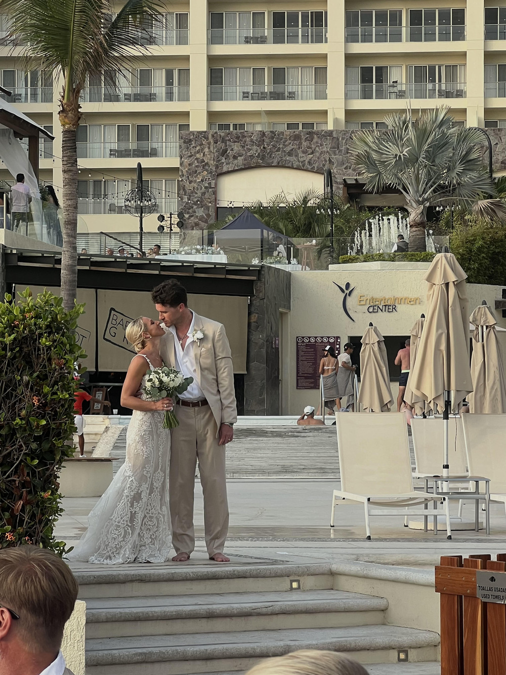 Leah and Christopher share a kiss outdoors at their destination wedding, bouquet in hand, with pool chairs from the resort visible in the background.