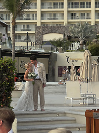 Leah and Christopher share a kiss outdoors at their destination wedding, bouquet in hand, with pool chairs from the resort visible in the background.