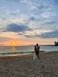Leah and Christopher stand hand in hand on a sandy beach at sunset, the city skyline glowing in the distance—a breathtaking backdrop for their destination wedding.