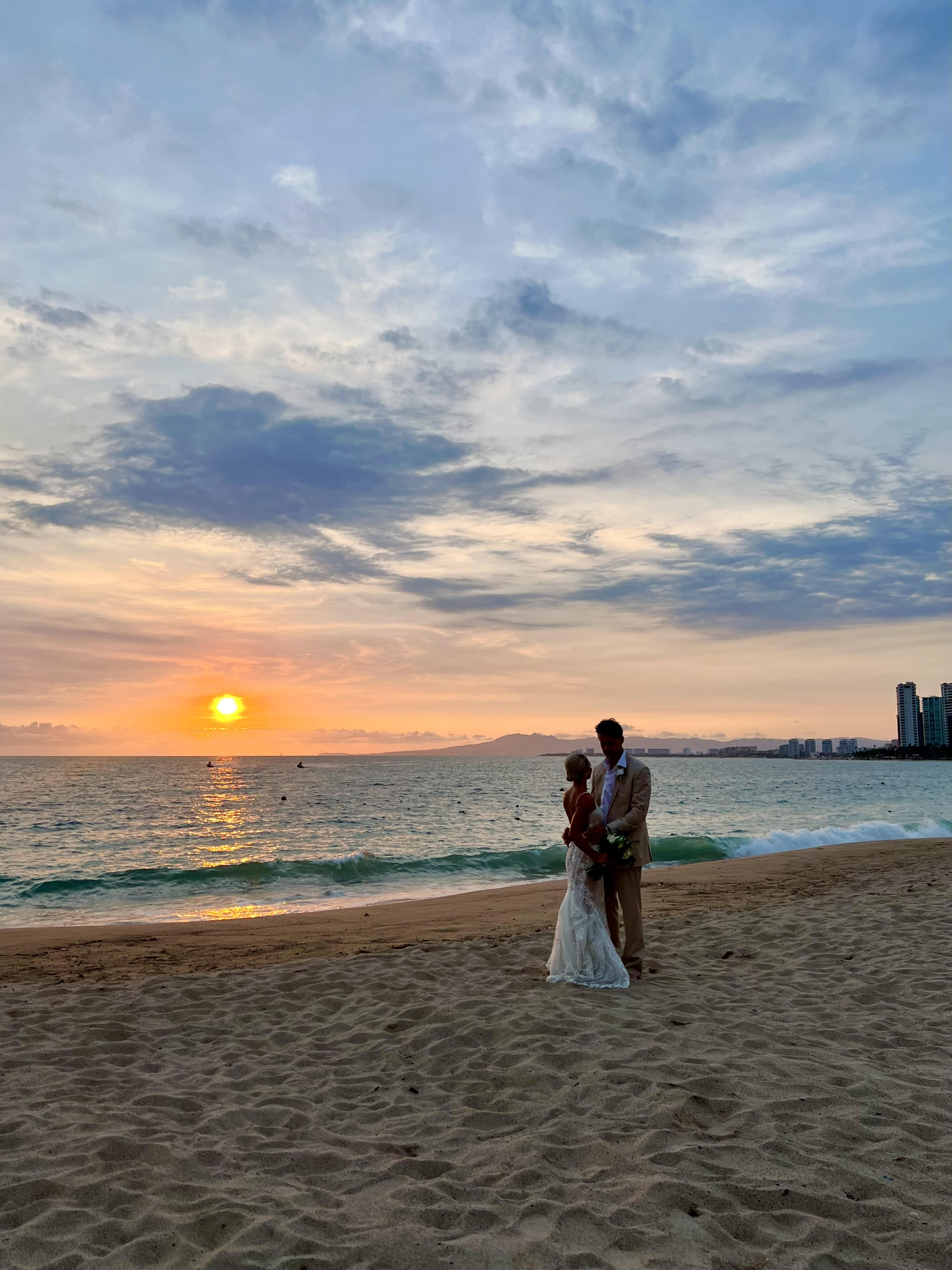 Leah and Christopher stand hand in hand on a sandy beach at sunset, the city skyline glowing in the distance—a breathtaking backdrop for their destination wedding.