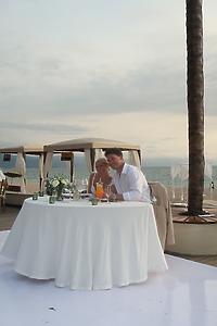 Leah and Christopher sit at a round dinner table by the beach, surrounded by cabanas, enjoying their destination wedding celebration.