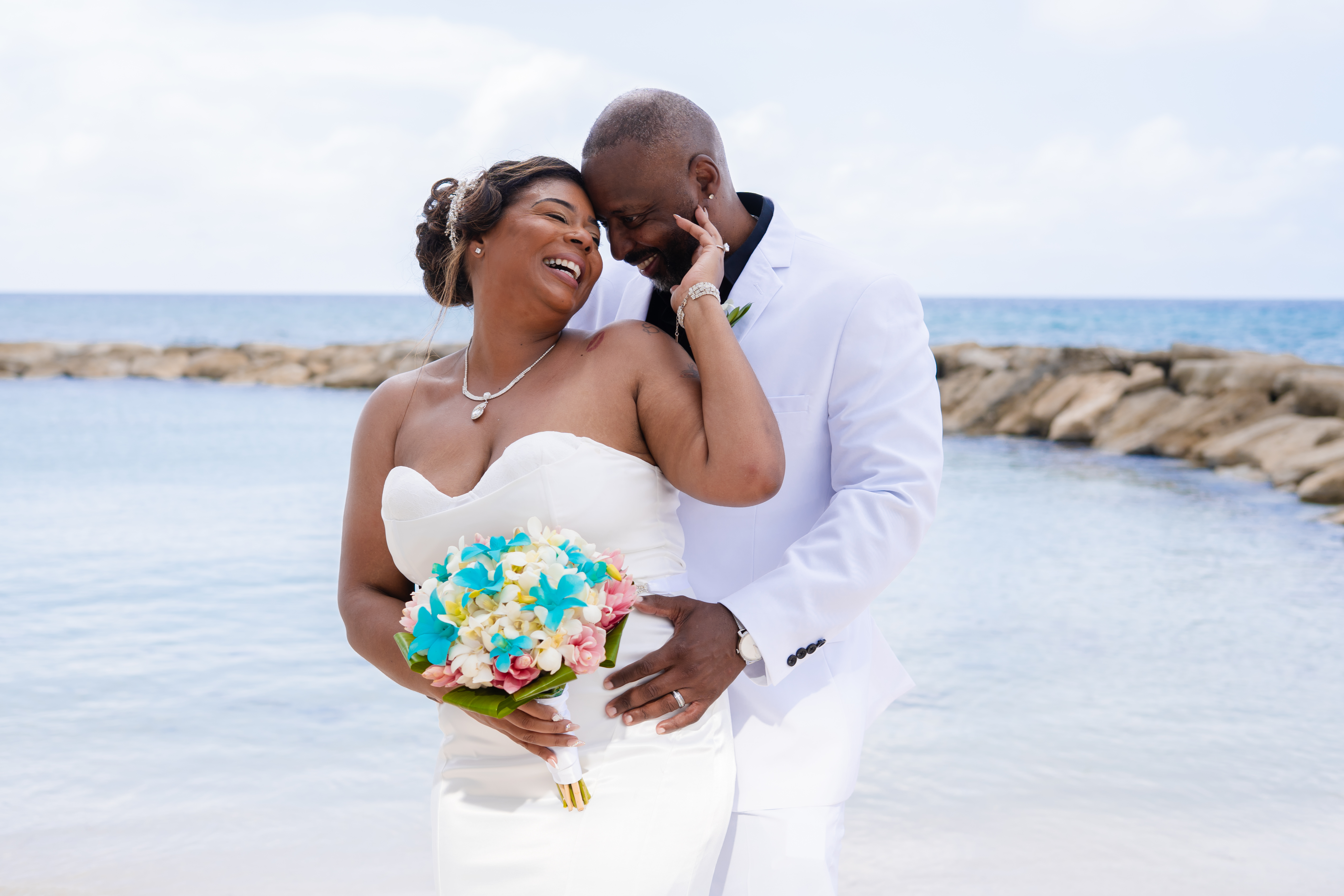 Kishan and Ericka stand by the water, smiling and embracing in their white wedding attire, capturing a perfect moment from their destination wedding.