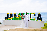 Kishan and Ericka stand in front of a large, colorful Jamaica sign by the ocean, celebrating their destination wedding.