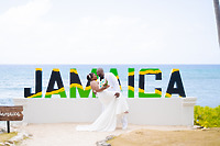 Kishan and Ericka pose in front of a large "JAMAICA" sign by the ocean, capturing a beautiful moment from their destination wedding.