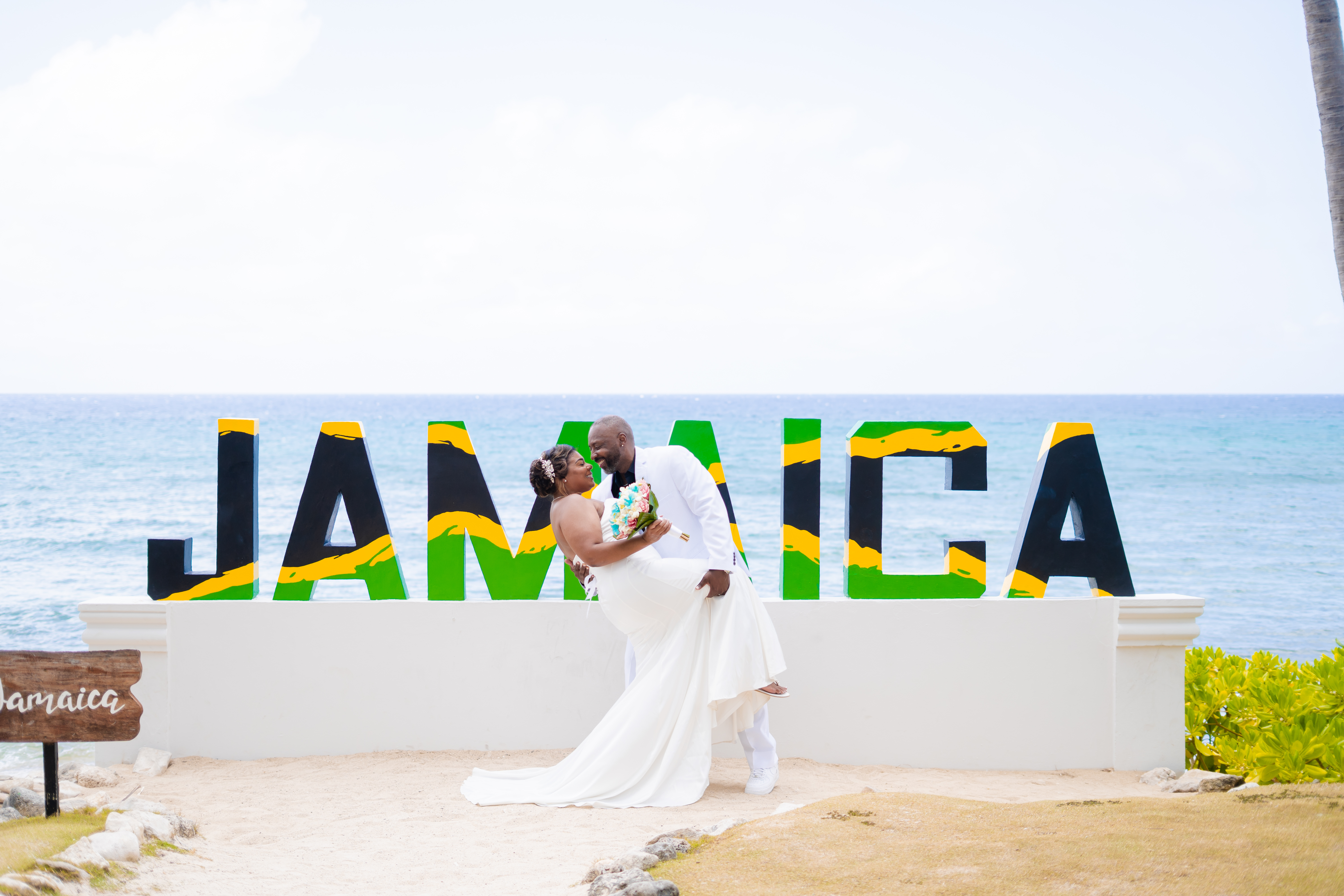 Kishan and Ericka pose in front of a large "JAMAICA" sign by the ocean, capturing a beautiful moment from their destination wedding.