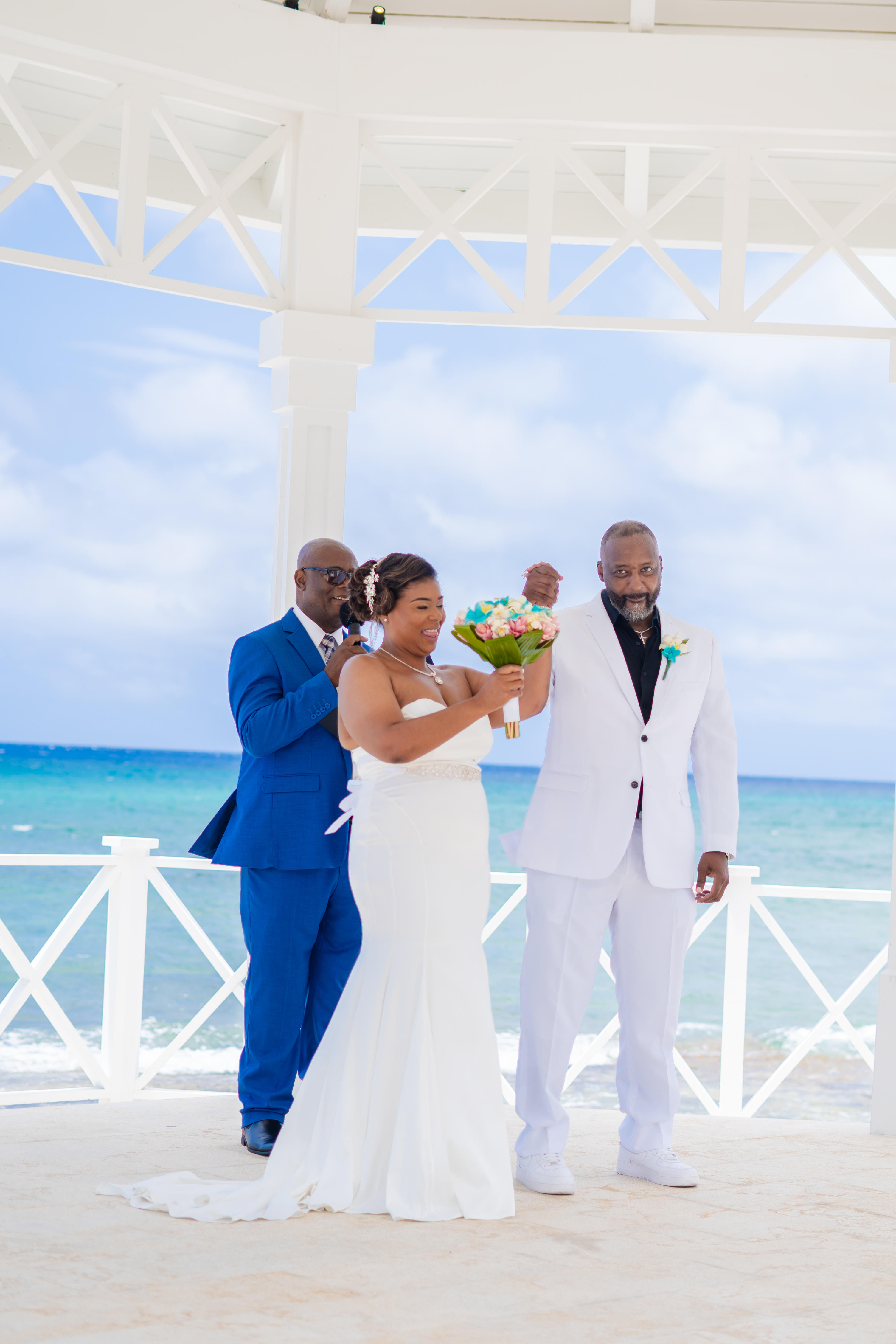 Kishan and Ericka stand under a gazebo by the ocean during their destination wedding, with a man in a blue suit positioned behind them.