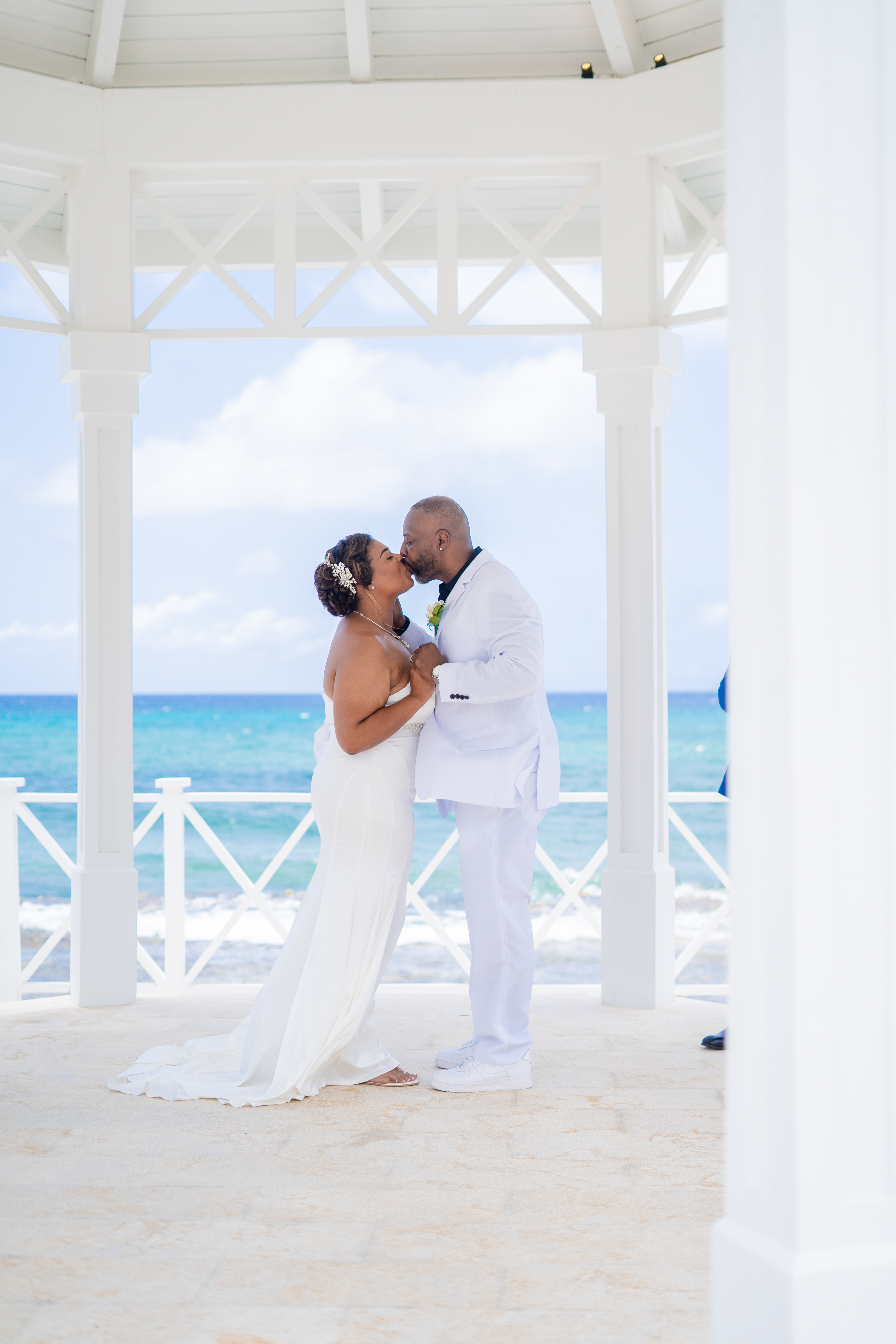Kishan and Ericka share a kiss beneath a white gazebo by the ocean, dressed in elegant wedding attire, capturing a beautiful moment from their destination wedding.