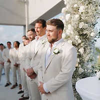 Trey and Kimberly’s groomsmen, dressed in light suits, stand beside a white floral display at their beautiful outdoor destination wedding.