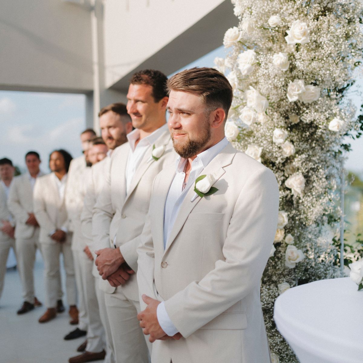 Trey and Kimberly’s groomsmen, dressed in light suits, stand beside a white floral display at their beautiful outdoor destination wedding.