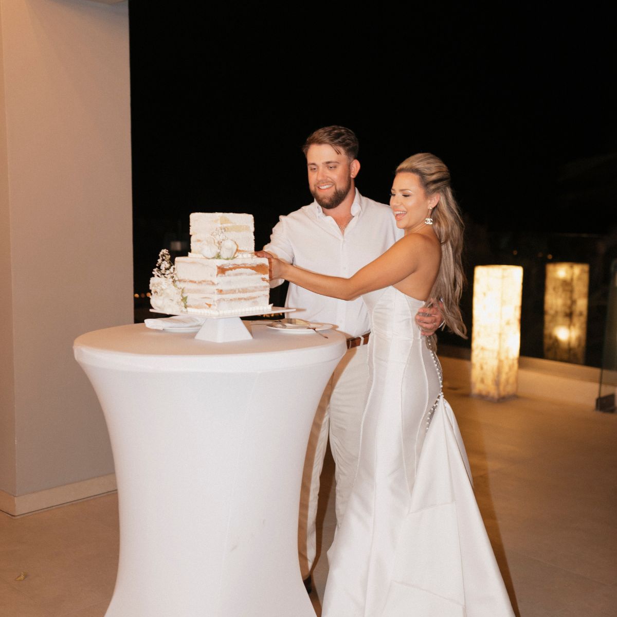 Trey and Kimberly, elegantly dressed in formal attire, cut a white tiered cake during their indoor destination wedding celebration.