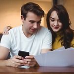 A couple sits together at a table, reviewing wedding documents; the man holds a smartphone as they plan their destination wedding.