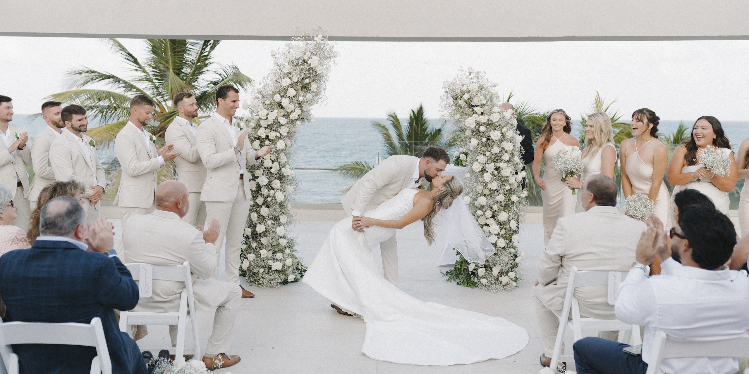 The bride and groom share a kiss during their outdoor destination wedding ceremony as guests applaud, with the ocean providing a stunning backdrop.