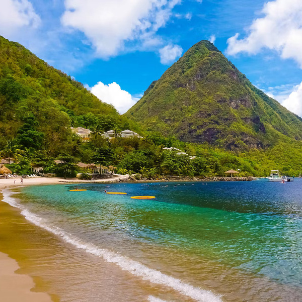 A perfect destination wedding spot, featuring a sandy beach with clear blue water, lush green hills, and a tall mountain rising in the background.