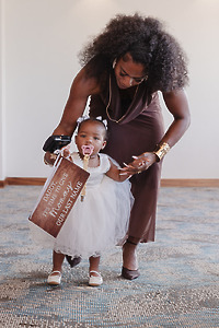 A woman helps a toddler in a white dress—adorned with Jordan and Kievan signs and holding a pacifier—take her first steps indoors at the destination wedding.