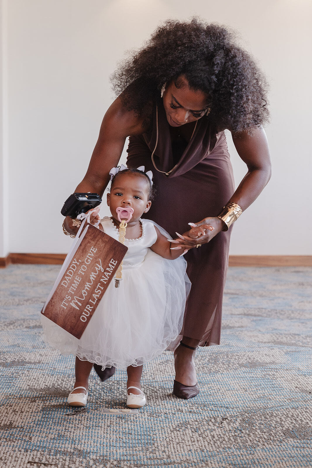 A woman helps a toddler in a white dress—adorned with Jordan and Kievan signs and holding a pacifier—take her first steps indoors at the destination wedding.