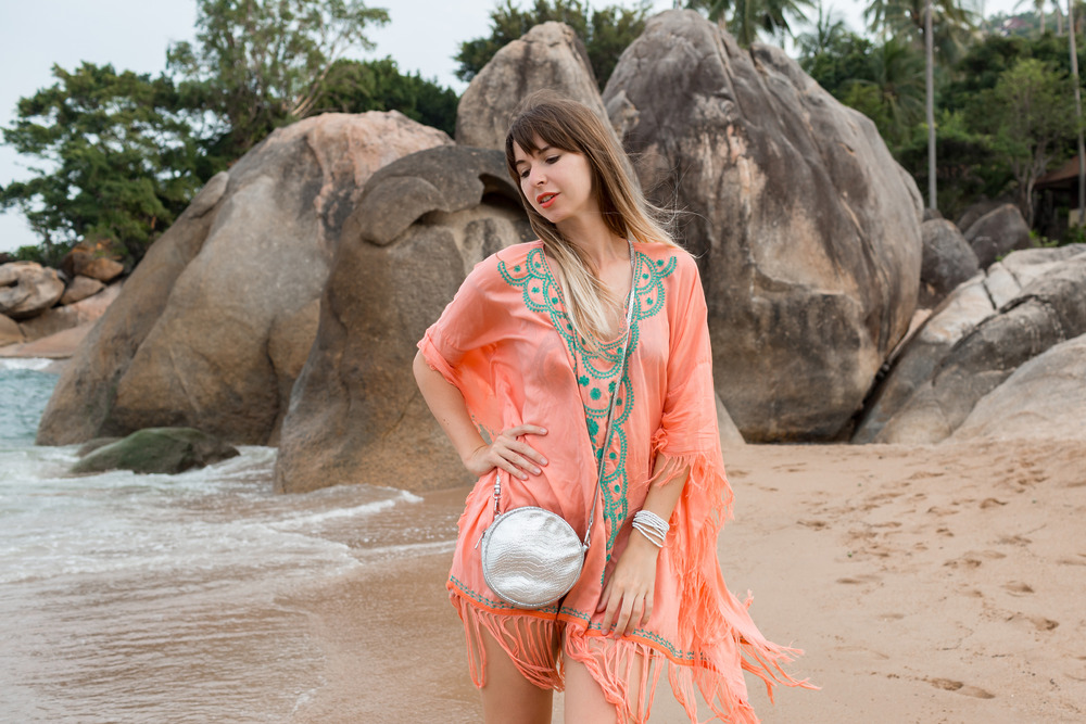 A woman in a peach beach cover-up stands on the sandy shore, with large rocks in the background—a perfect setting for a destination wedding.