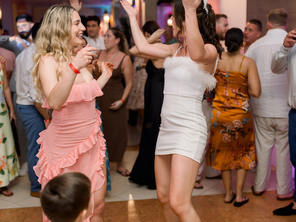 Two women in dresses dance and smile together at a crowded indoor destination wedding celebration.