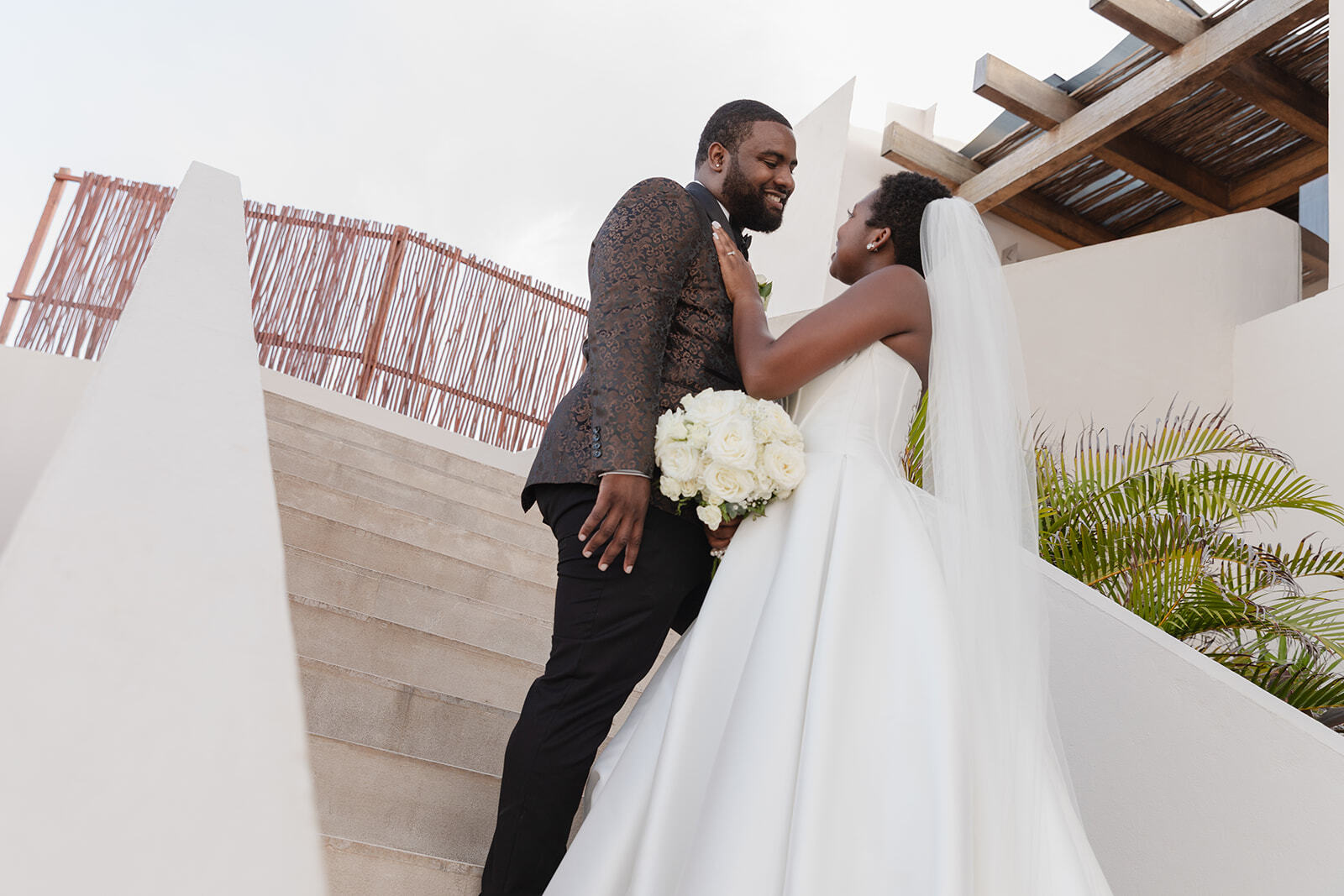 Saphire and Stephen pose together on outdoor stairs during their destination wedding, with the bride holding a bouquet of white roses.