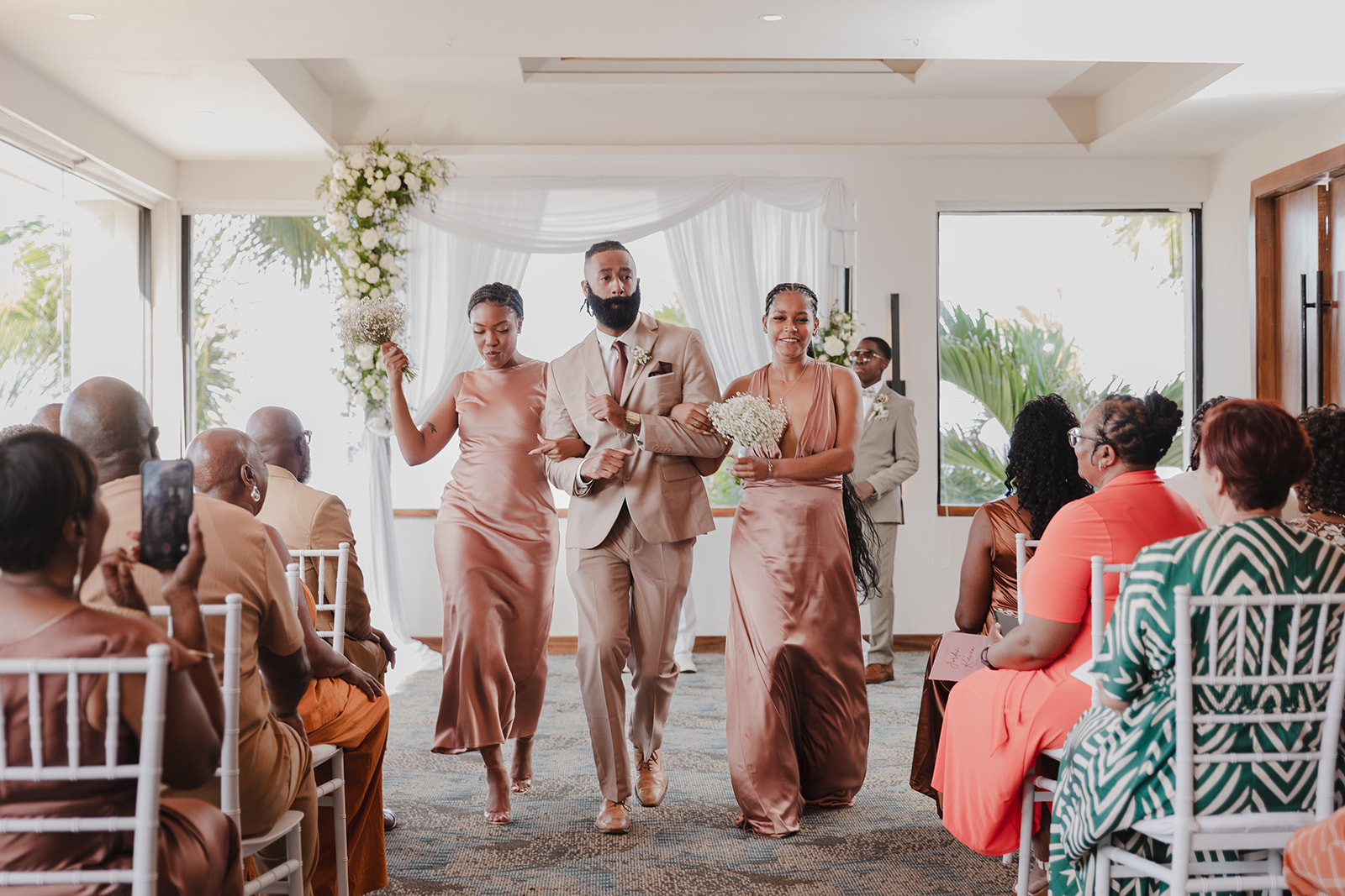 Jordan and Kievan walk down the aisle with a third attendant as wedding guests watch in a bright room.