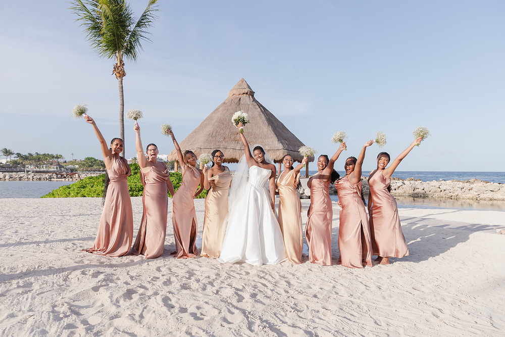 Bride and bridesmaids in Jordan and Kievan gowns with bouquets at a sunny destination wedding beach.