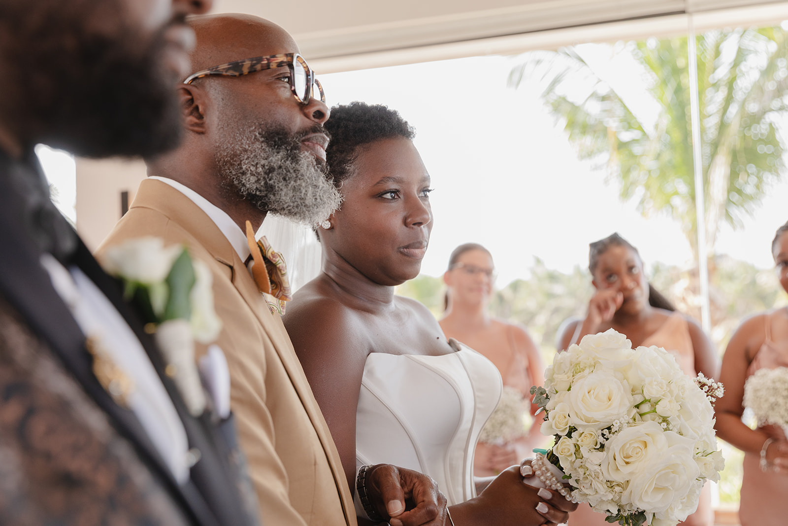 At a destination wedding, the bride holding a bouquet stands next to a man in a tan suit.