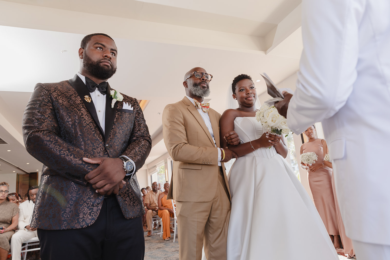 At the destination wedding, Jordan and Kievan stand indoors as the bride holds a bouquet by her escort.
