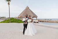 Saphire and Stephen pose with their young child on a sandy beach near a thatched-roof hut, capturing a special moment during their destination wedding.