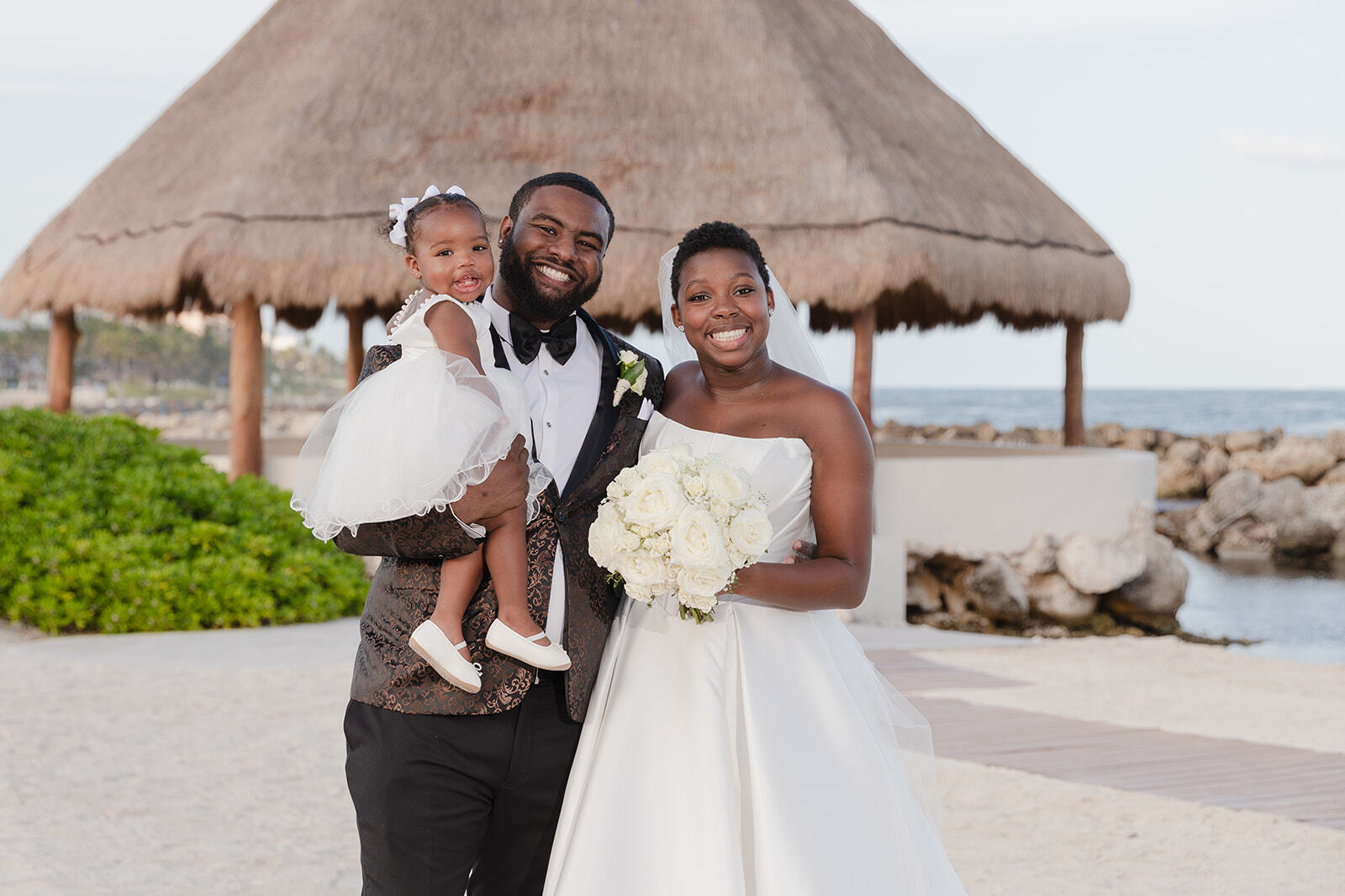 Saphire and Stephen smile and pose on the beach with a young girl, a rustic thatched hut in the background, capturing a joyful moment at their destination wedding.