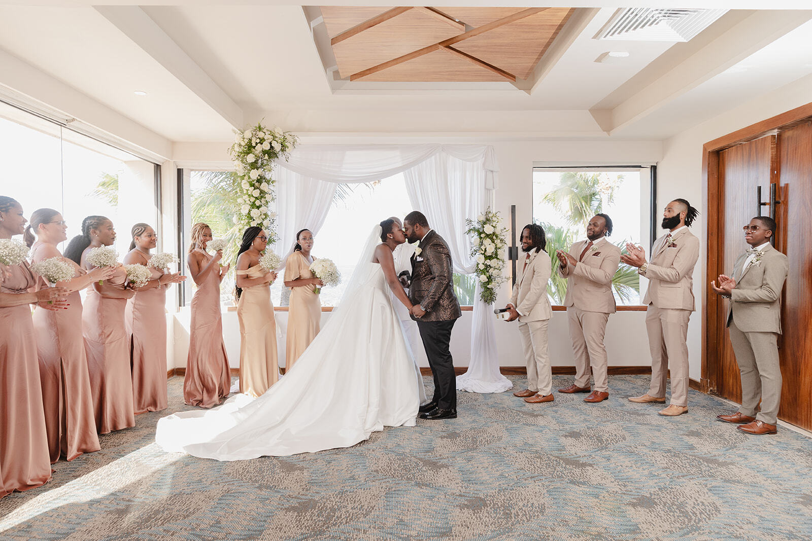 Saphire and Stephen share a kiss at their beautiful indoor wedding, surrounded by their bridesmaids and groomsmen.