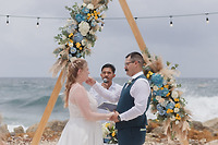 Teneal and Jonathan exchange vows during a stunning destination wedding ceremony on the beach, standing beneath a floral arch with an officiant presiding.