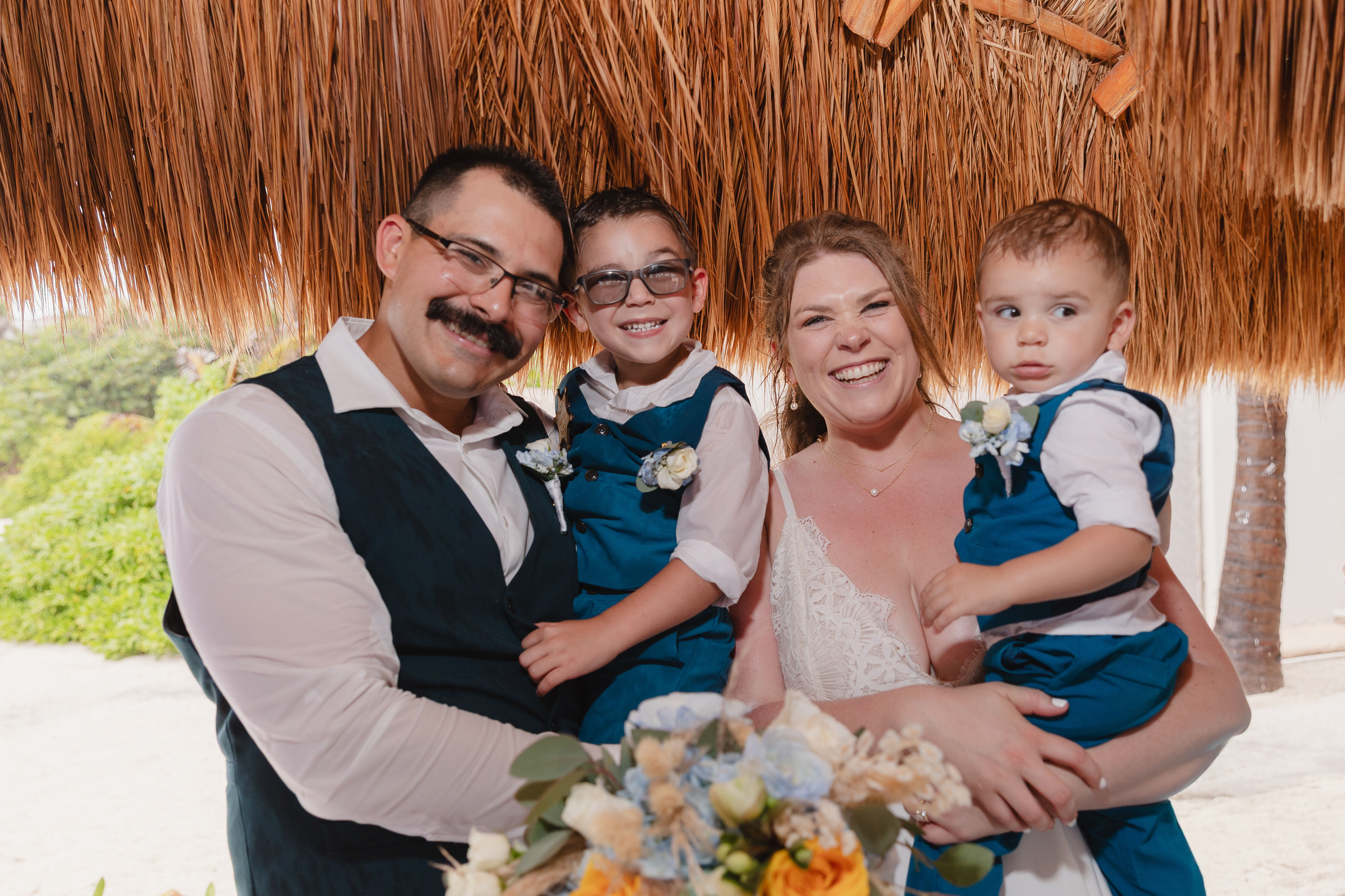 A family of four poses and smiles beneath the thatched roof at Teneal and Jonathan's destination wedding.