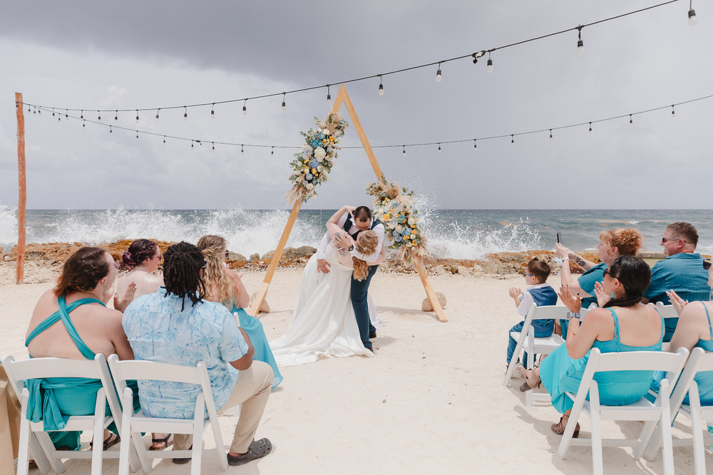 Teneal and Jonathan share a kiss at their beautiful destination wedding on the beach, surrounded by guests who watch and capture the special moment with photos.