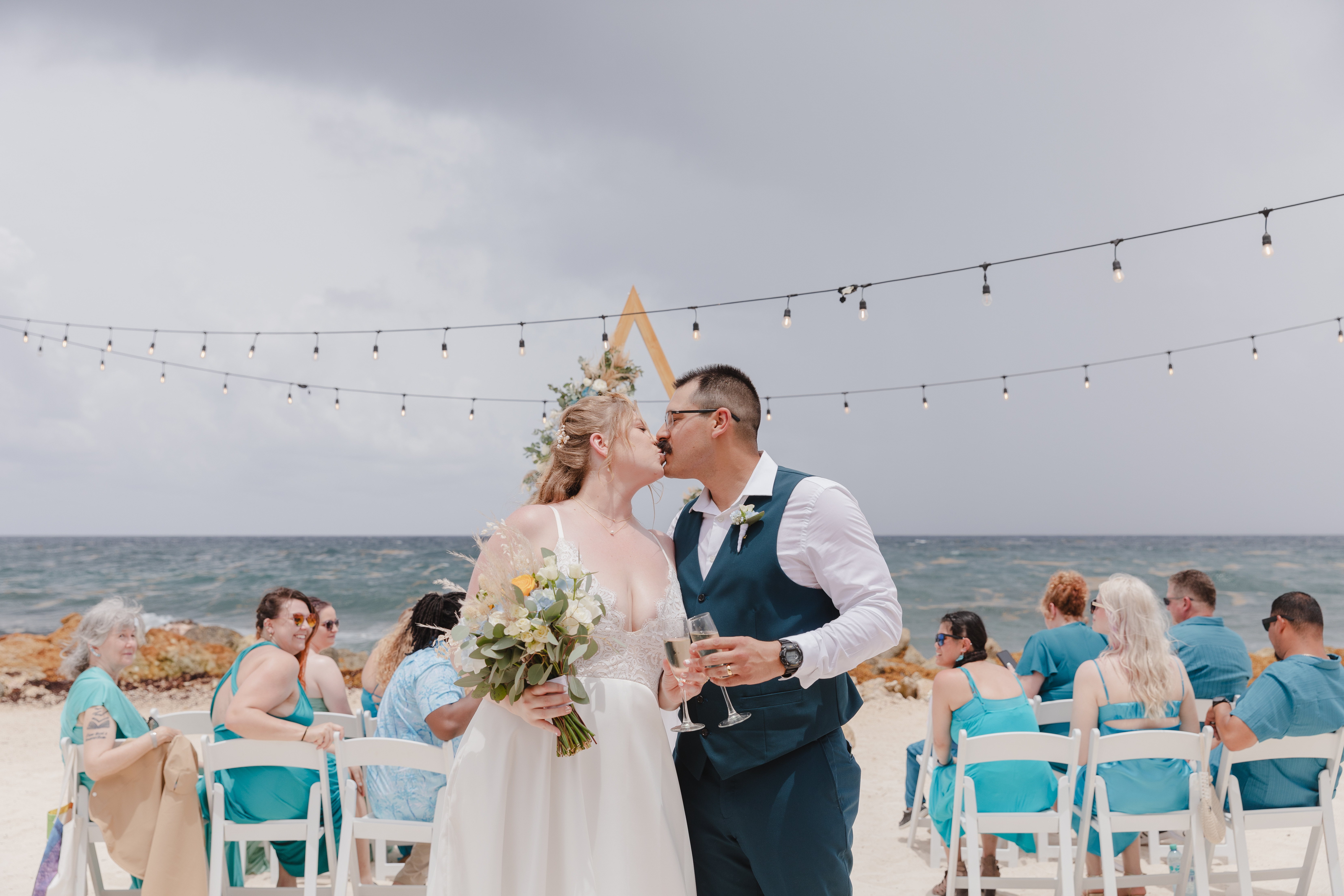 Teneal and Jonathan share a kiss during their destination wedding ceremony on the beach, with their guests seated behind them, witnessing the special moment.