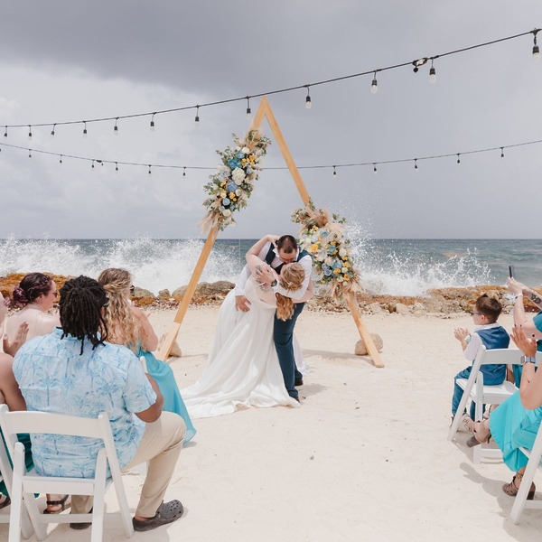 Teneal and Jonathan share a romantic kiss during their destination wedding on the beach, surrounded by loved ones and the sound of waves in the background.