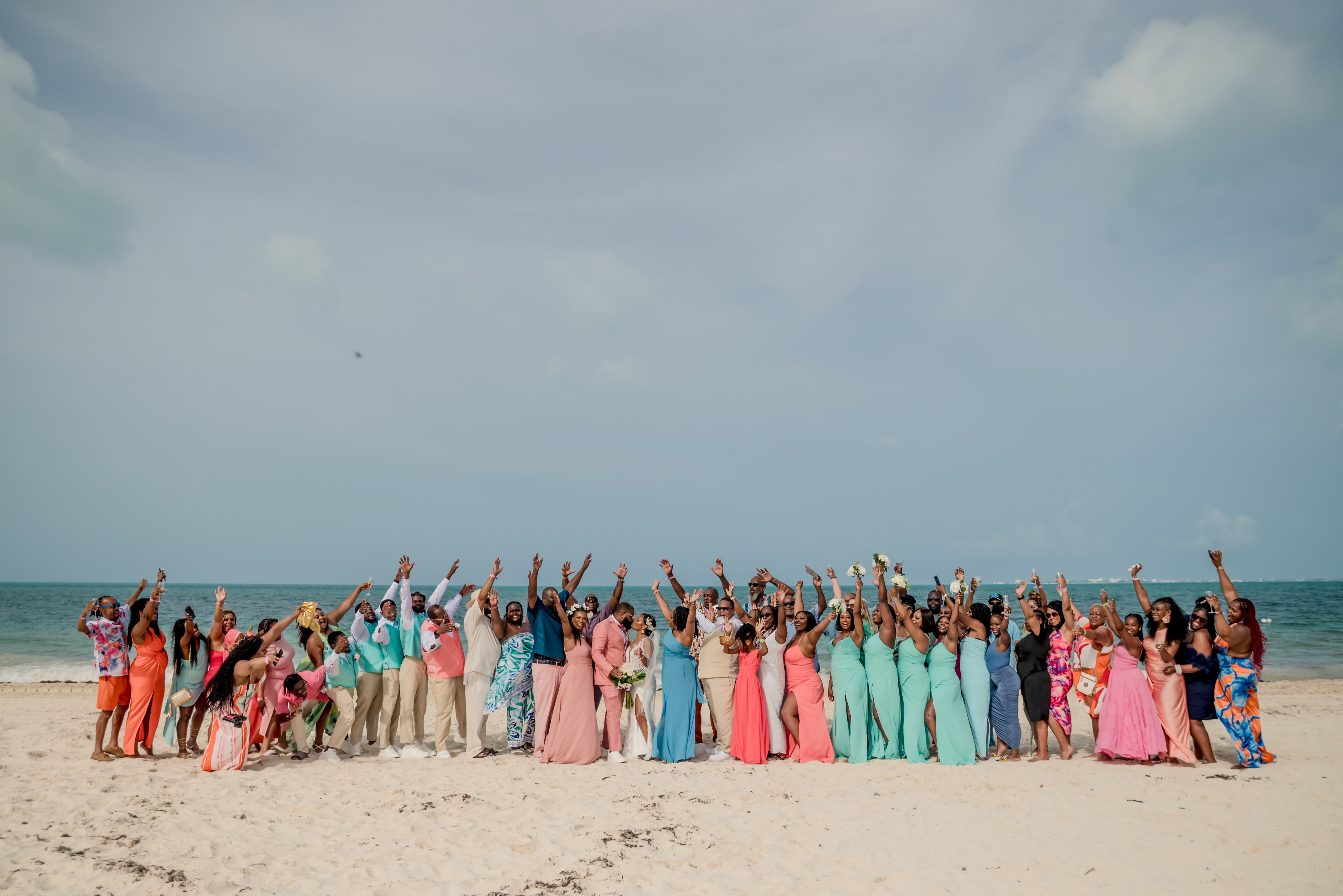 A large group gathers and cheers on a sandy beach, celebrating Sydney and Hayden's destination wedding.
